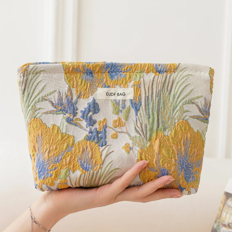 A hand holds a floral canvas makeup bag featuring blue and yellow flowers and green leaves.