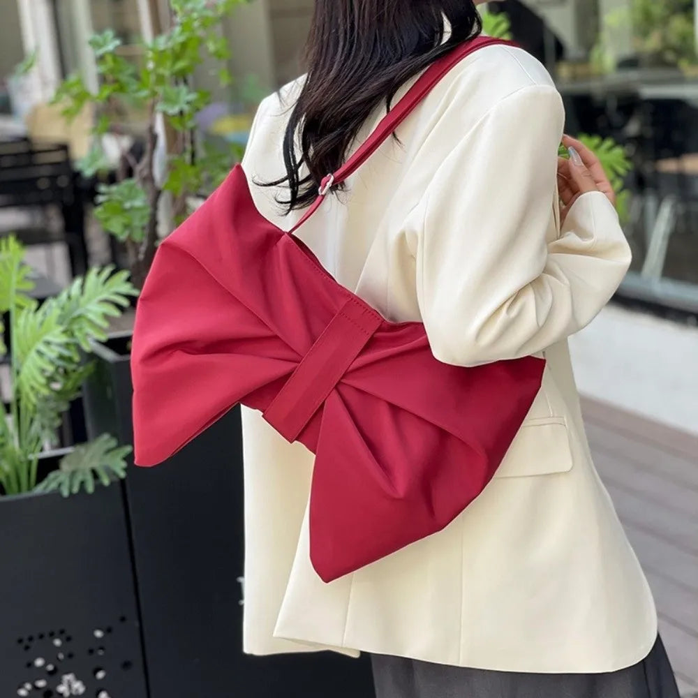 A woman holds a red pleated bow crossbody tote bag against a light colored blazer outdoors.