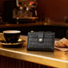 A black leather square folding wallet rests on a wooden counter next to a cup of coffee.