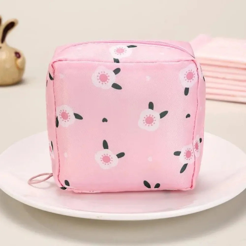 A pink fabric pouch designed for storing sanitary napkins with a white floral pattern, resting on a white plate.