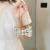 A hand holds a silver heart-shaped acrylic evening clutch with a chain strap against a light background.