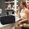 A woman holds a black travel toiletry bag while sitting at a counter in a well-lit room.