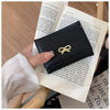 Cartera de cuero PU negra tríptico con cierre trenzado dorado, sostenida por una mano contra un fondo de papel impreso.