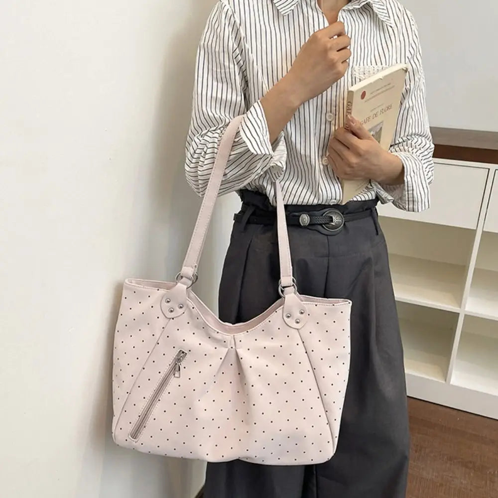 A woman holds a light pink retro polka dot shoulder tote bag against a white wall.