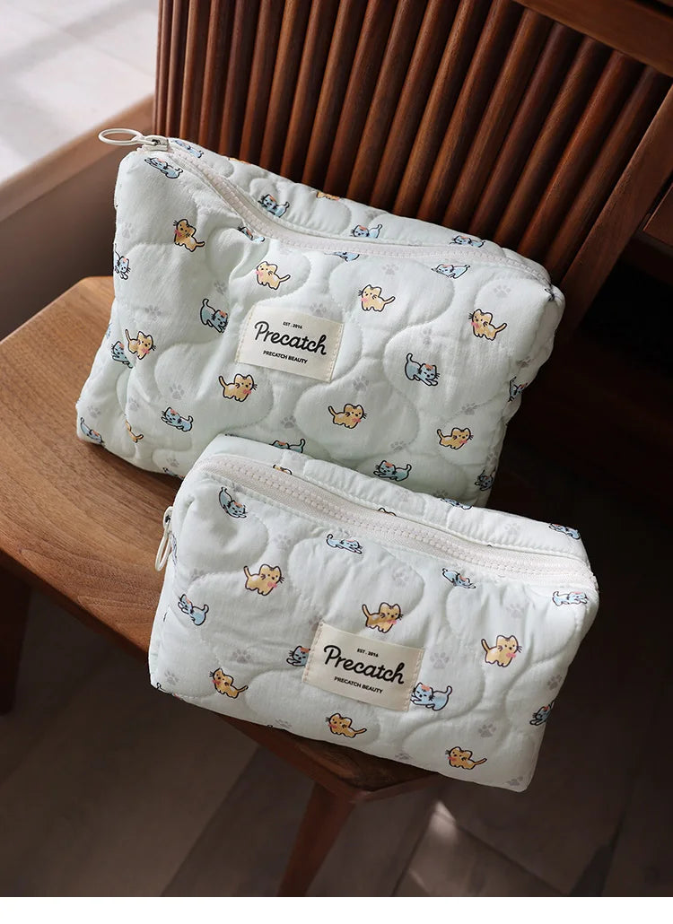A white quilted cosmetic storage pouch with a light blue pattern of small cats resting on a wooden surface.