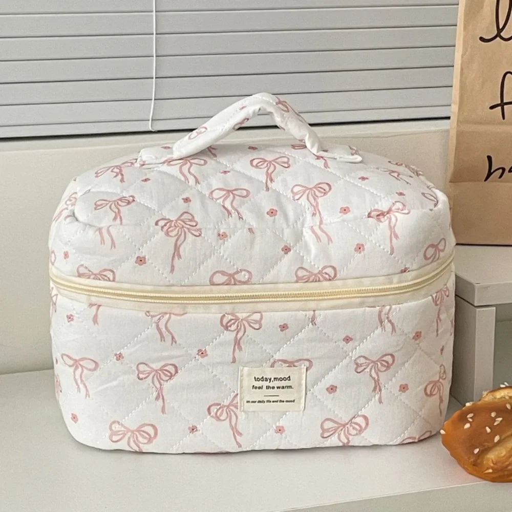 A white quilted cosmetic bag with a pink floral pattern and a light yellow zipper, resting on a white surface.