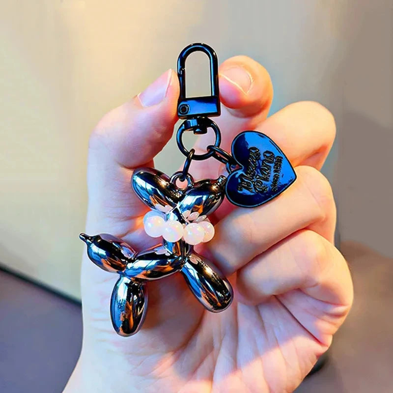 A hand holds a shiny metallic balloon dog keychain charm with a hook and pearl accents.