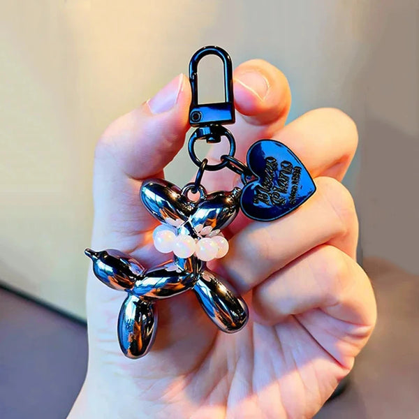 A hand holds a shiny metallic balloon dog keychain charm with a hook and pearl accents.