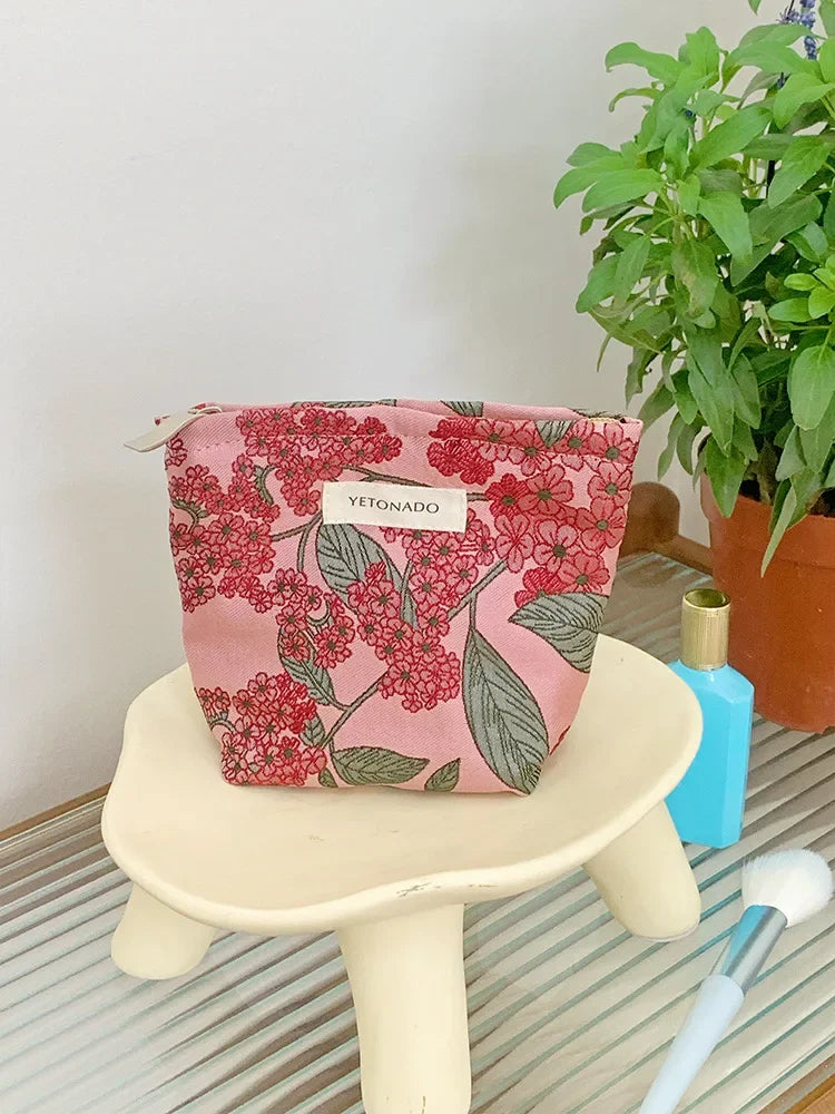 A pink floral patterned cosmetic pouch resting on a light-colored stool next to a potted green plant.