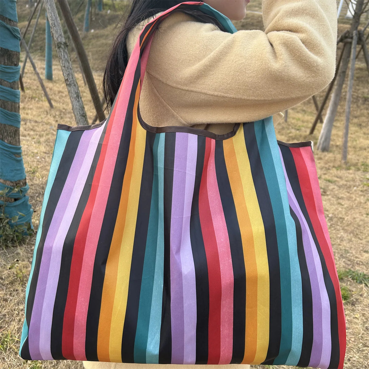 A person holds a colorful striped grocery tote bag with vertical stripes of various colors outdoors.