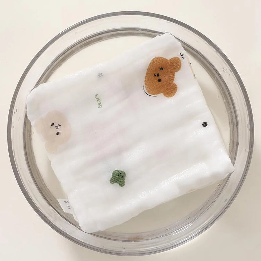 A white, multi-layered cotton baby burp cloth towel with small bear-shaped appliqués is shown in a clear glass bowl.
