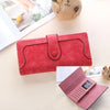 A red leather clutch wallet is shown next to a pink version, with a small stuffed animal in the background.