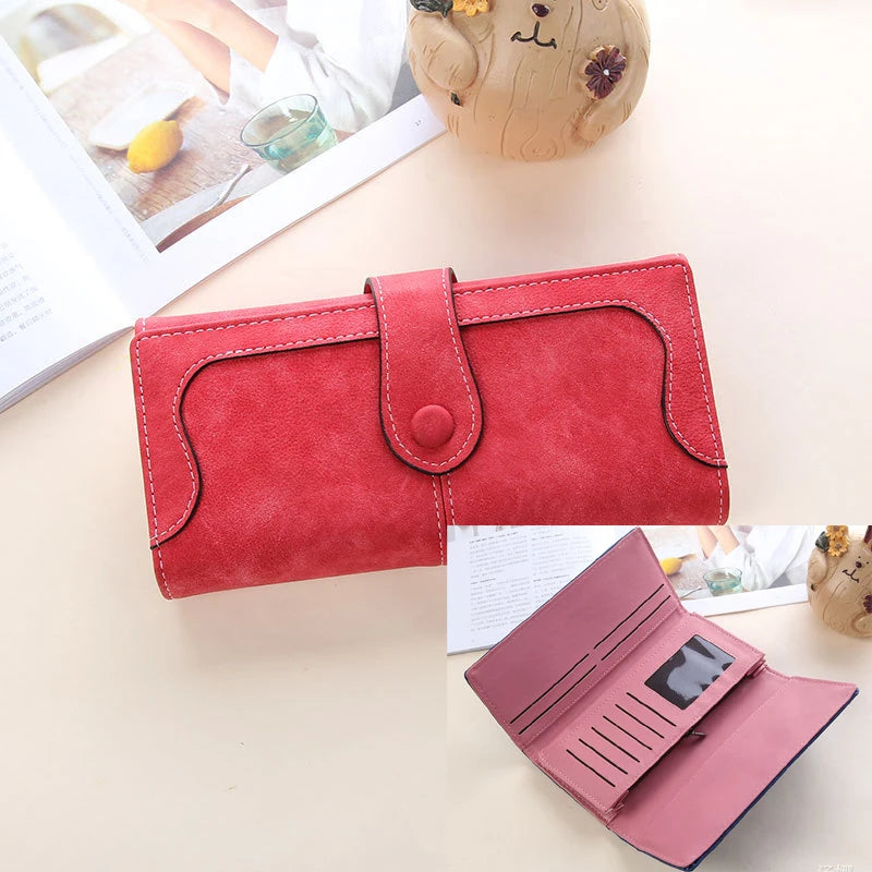 A red leather clutch wallet is shown next to a pink version, with a small stuffed animal in the background.