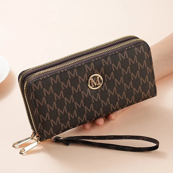 A dark brown monogrammed double zipper wallet with a gold logo and leather strap resting on a light background.