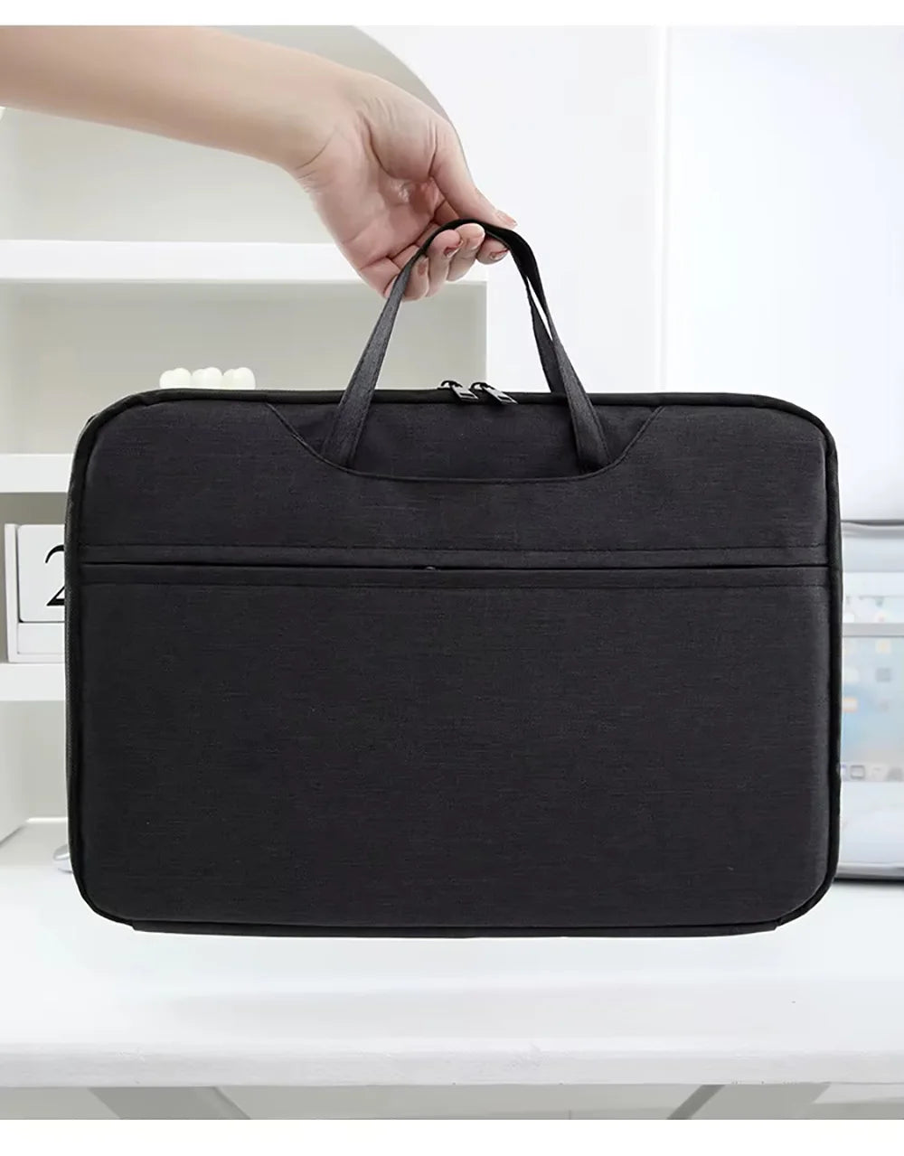 A black laptop sleeve with a handle is being held by a hand, shown on a white surface.