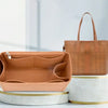A tan felt insert liner for a handbag is displayed next to a brown leather tote bag and a white cylindrical container.
