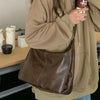 A person wearing a beige hoodie holds a brown vintage PU leather tote bag with a crossbody strap.