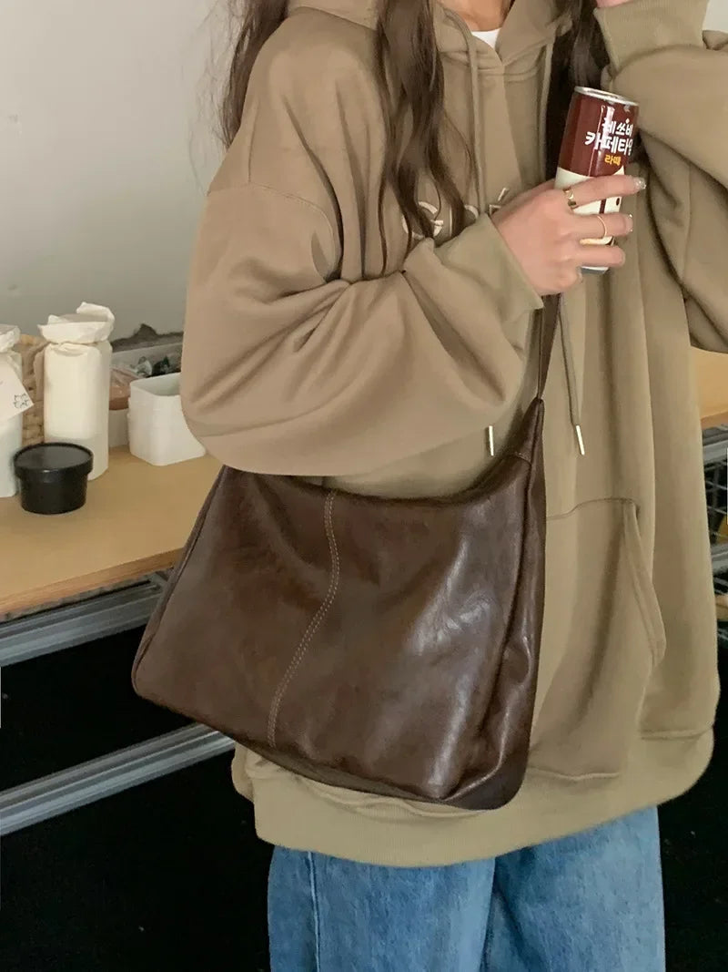 A person wearing a beige hoodie holds a brown vintage PU leather tote bag with a crossbody strap.