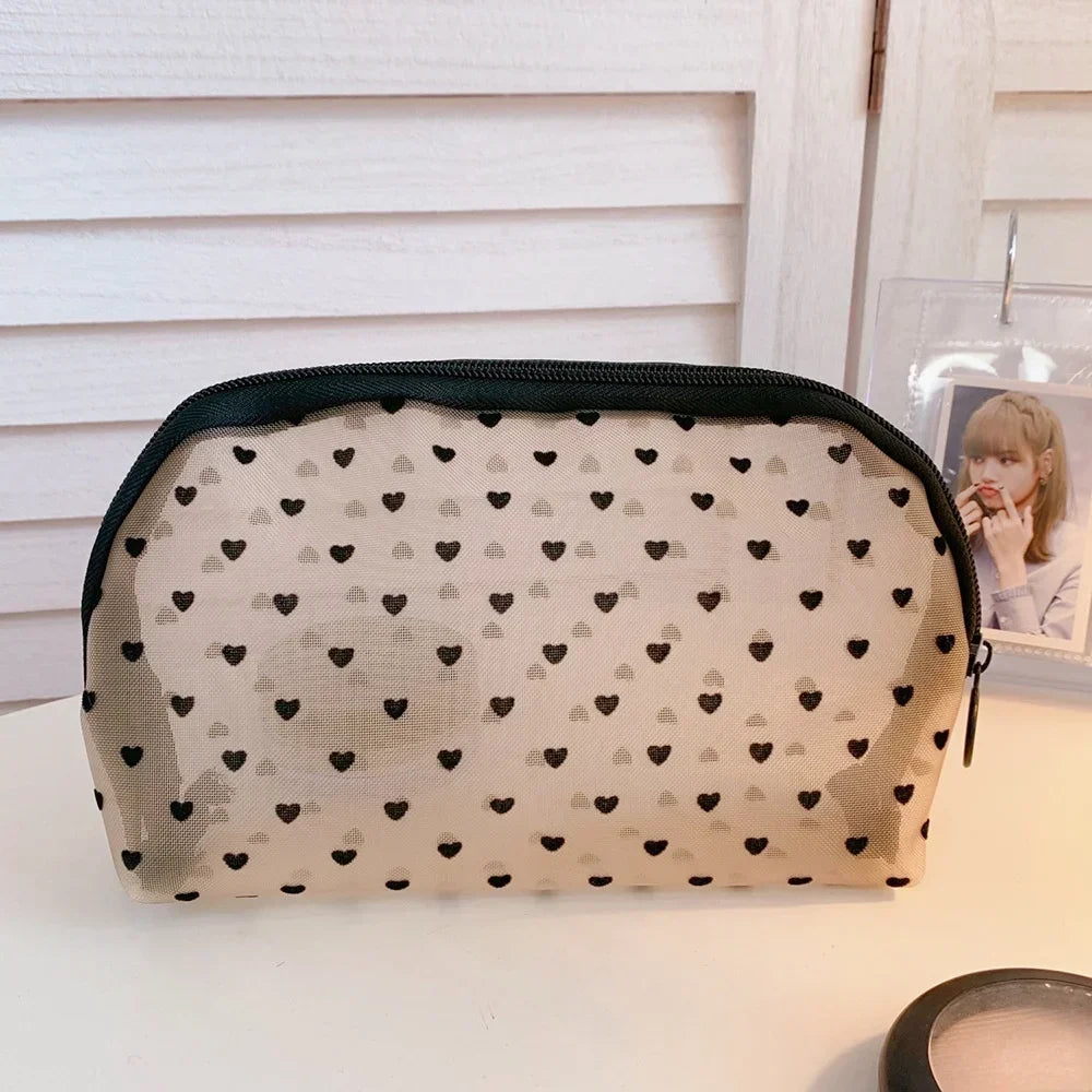 A beige mesh cosmetic storage pouch with a black trim and small black hearts pattern, resting on a white surface.