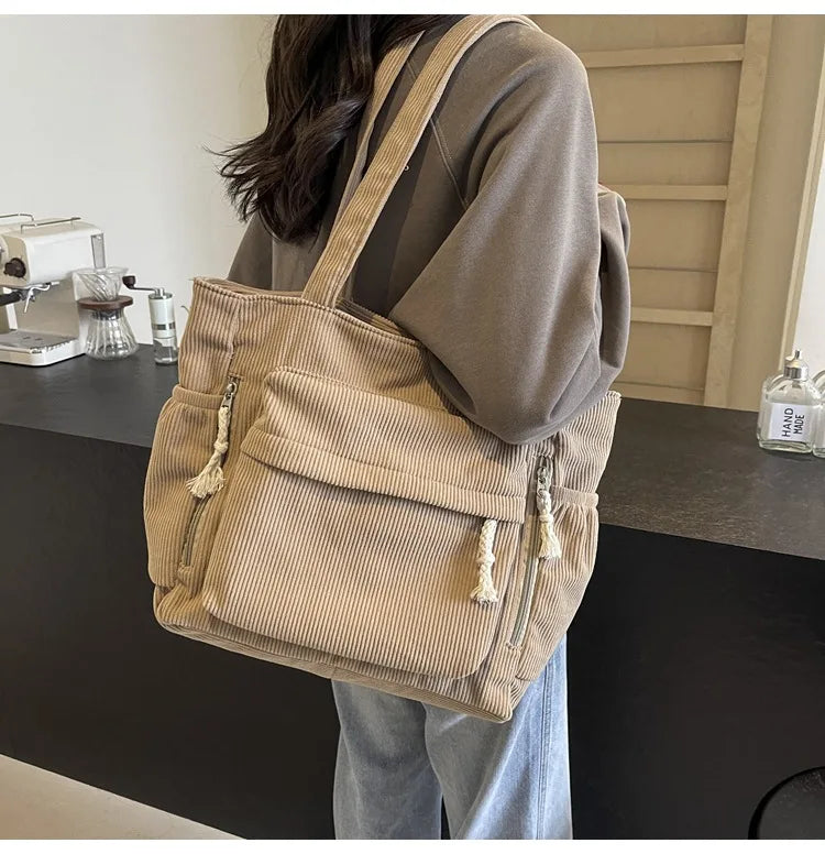 A beige corduroy shoulder bag with zipper details is shown hanging on a person's shoulder.