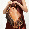 A woman holds a brown leather fringed clutch bag with tassels against a light background.