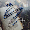 Blaue Rosenkranzkette mit silbernem Kreuzanhänger auf einem Textblatt.