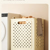 A light beige foldable laundry basket with a perforated design is shown on a floor, next to a white cabinet.