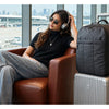 A woman is sitting in a leather chair with a large black laptop backpack next to her, in an airport-ready setting.