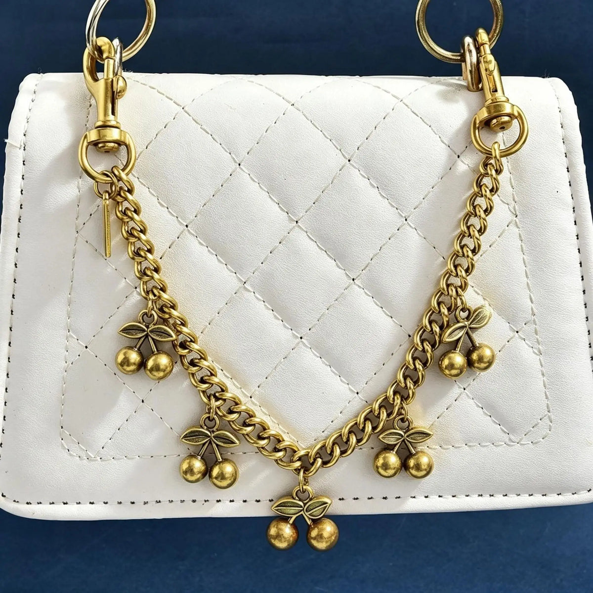 Collar de cadena de oro con adornos de cereza sobre un bolso de cuero blanco acolchado.