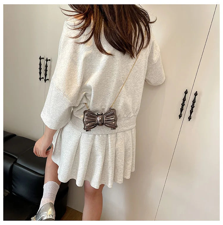 A woman models a light gray acrylic bow clasp mini handbag with a gold chain strap against a white wall.