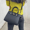 A black genuine leather shoulder bag carried by a person wearing a yellow sweater and dark pants.