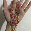 A hand holds a colorful resin bag strap necklace with pink and green hues and gold charms.