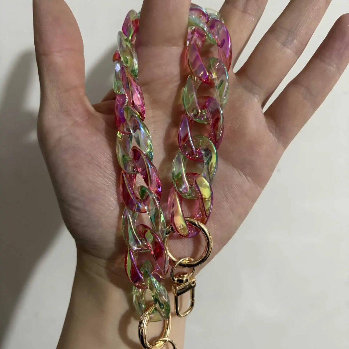 A hand holds a colorful resin bag strap necklace with pink and green hues and gold charms.