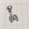 A silver stainless steel alphabet letter charm, the letter M, hanging from a lobster clasp, placed on a ruler.