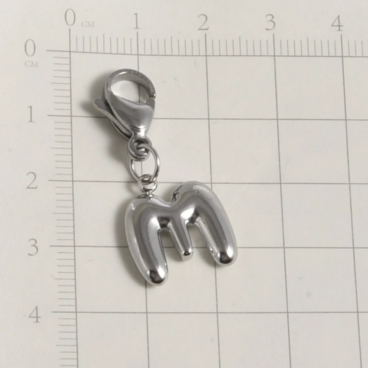 A silver stainless steel alphabet letter charm, the letter M, hanging from a lobster clasp, placed on a ruler.