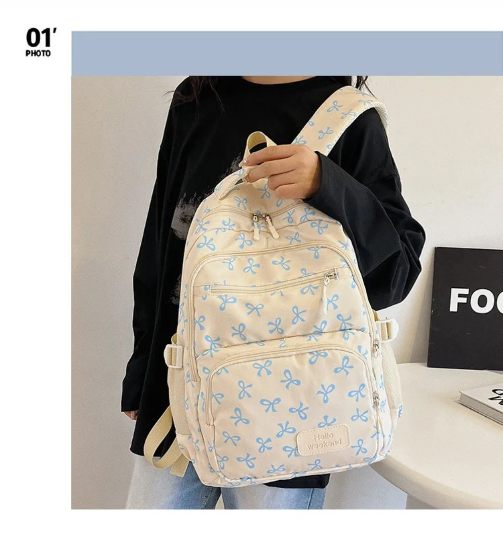 A light cream nylon backpack with a blue bow print is carried by a person against a white background.
