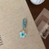 A light blue resin flower keychain with a silver jump ring and a blue metal clasp resting on a beige fabric surface.