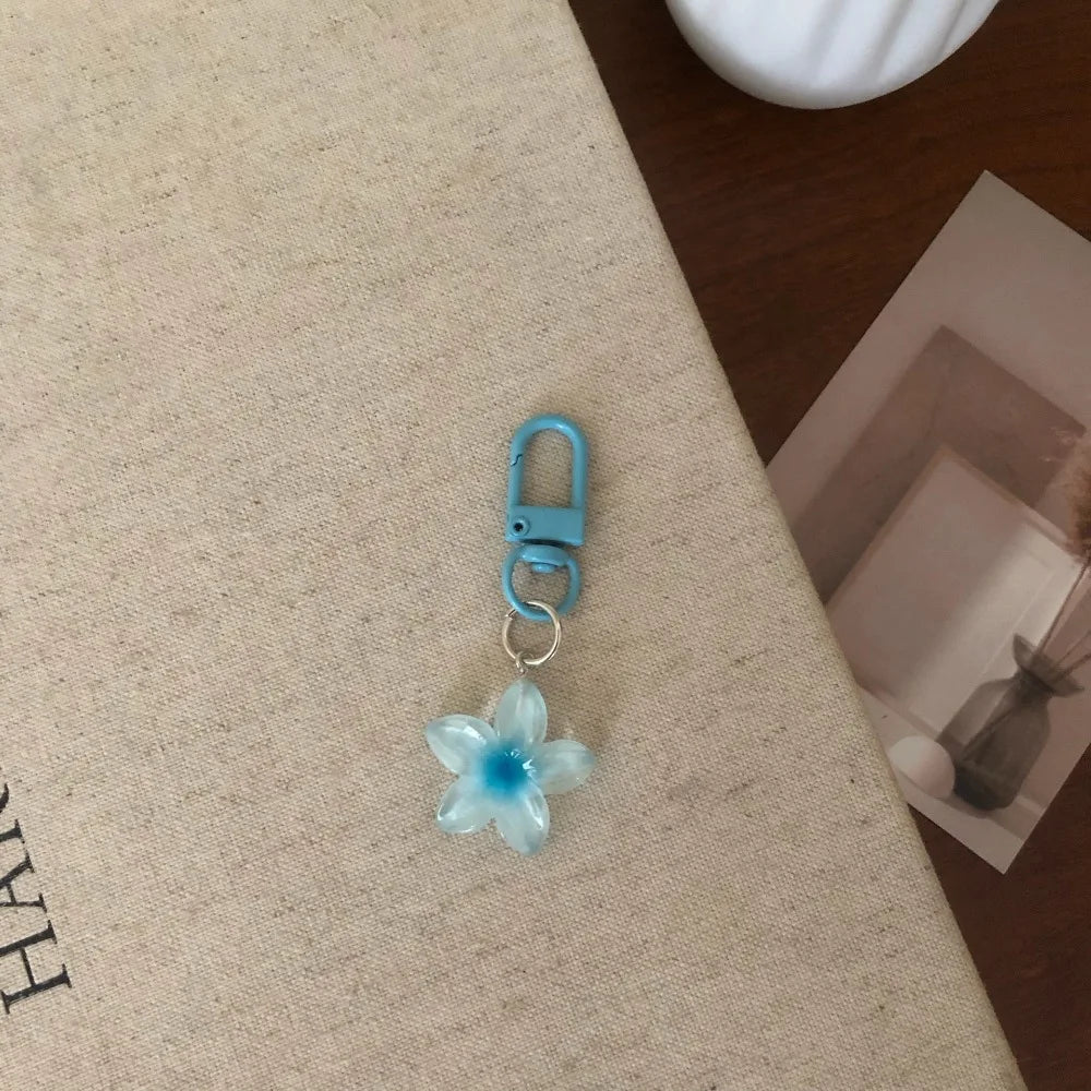 A light blue resin flower keychain with a silver jump ring and a blue metal clasp resting on a beige fabric surface.