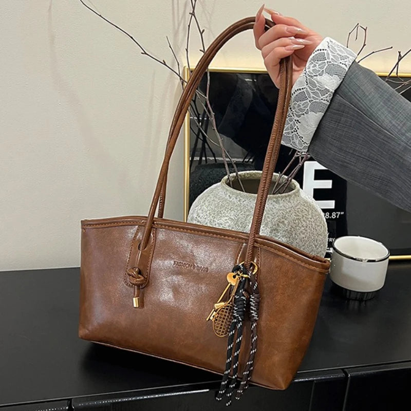 A brown classic PU leather tote bag with gold hardware is shown next to a white ceramic cup and dried branches.