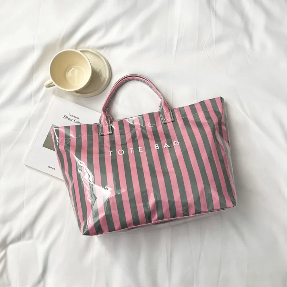A pink and black striped transparent PVC tote bag resting on a white sheet with a cup nearby.