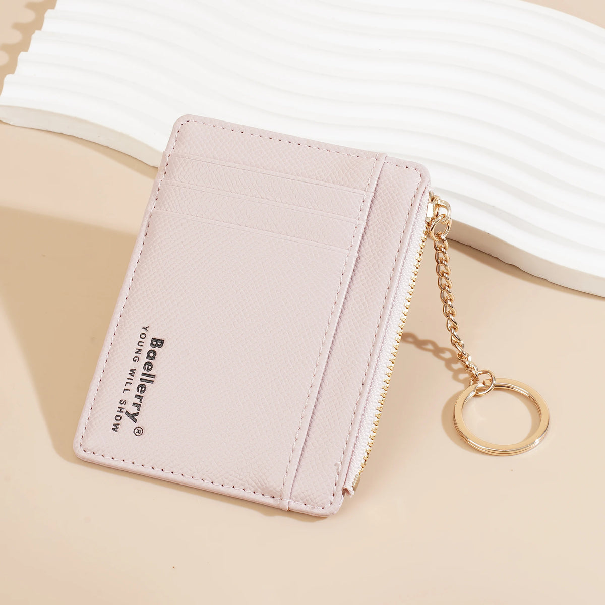 A pale pink ultra-thin card holder wallet with a gold chain is shown resting on a light beige surface.
