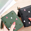 A hand holds a green PU leather wallet with small pink and white heart embellishments next to a black version.