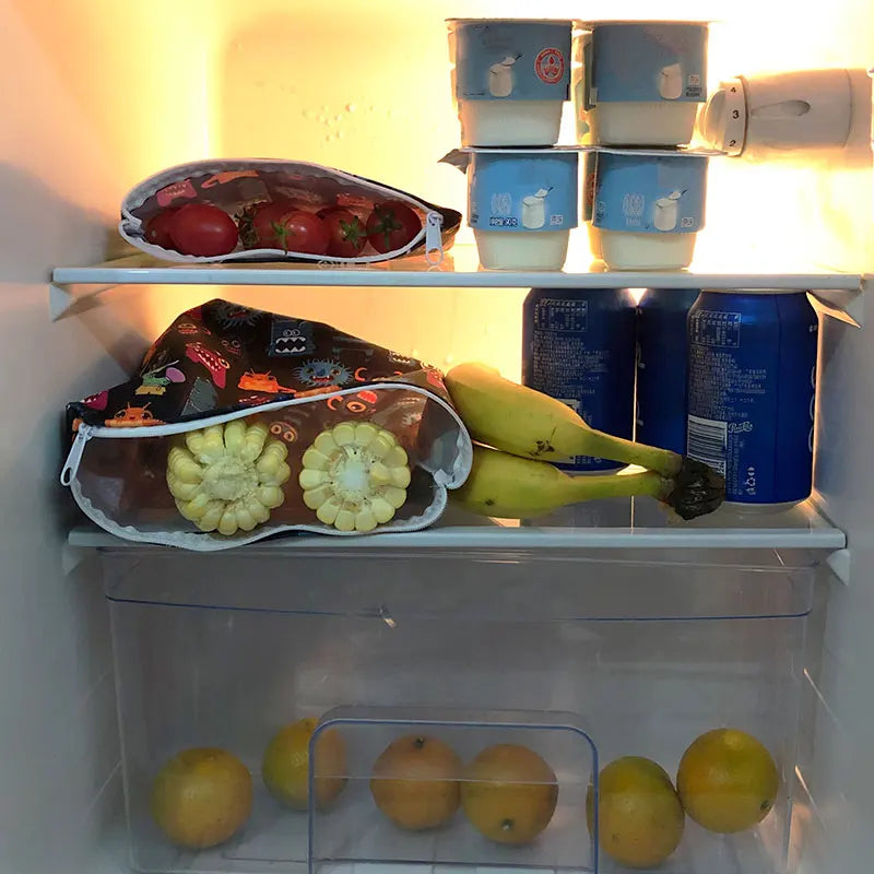 A display of reusable waterproof snack bags filled with fresh fruit like apples, tomatoes, and bananas.