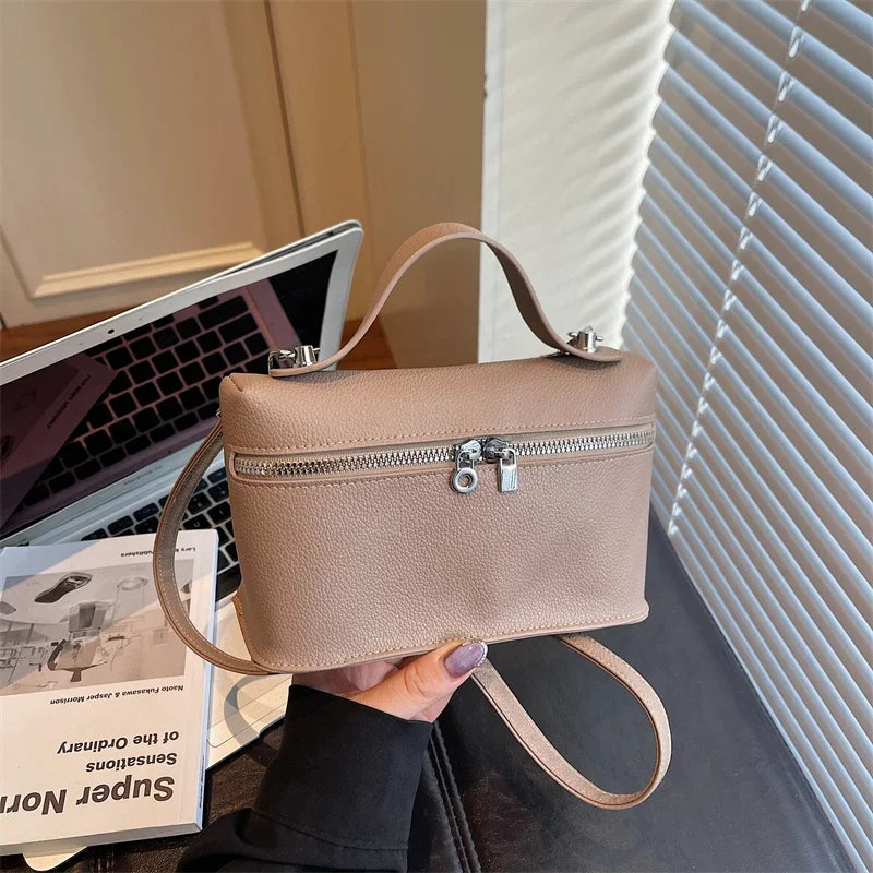 A light beige pebbled leather box crossbody bag with a tan leather strap is held against a white window blind.