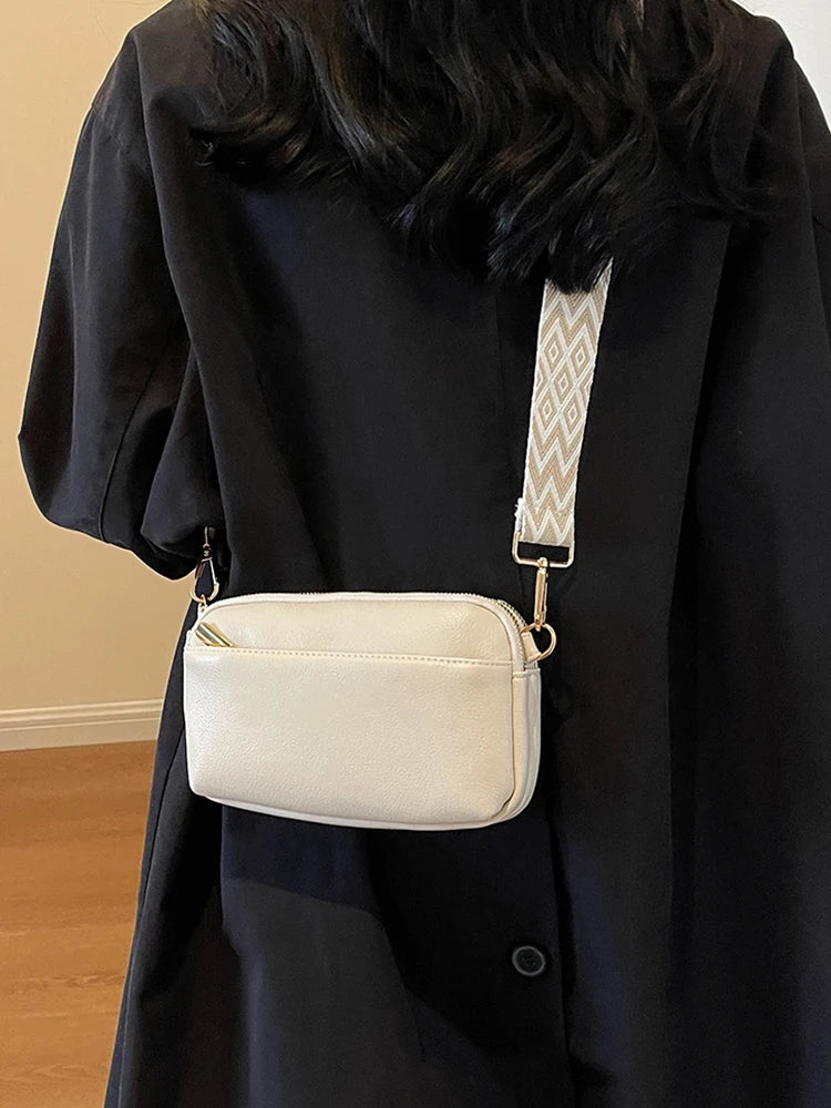 A cream-colored vegan PU crossbody bag with a geometric patterned strap is shown hanging from a black coat.