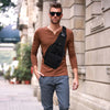 A man wearing a brown long-sleeved shirt and gray pants carries a black tactical sling backpack outdoors.