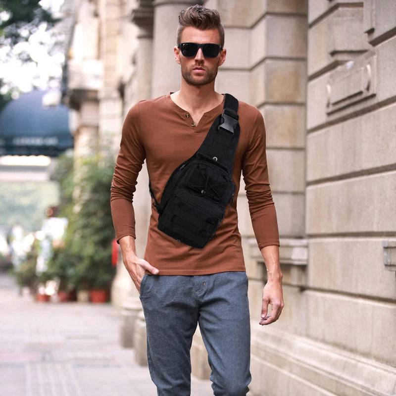 A man wearing a brown long-sleeved shirt and gray pants carries a black tactical sling backpack outdoors.