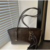 A brown leather tote bag with a long strap is displayed against a white slatted blind.
