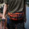 A person wearing a dark shirt and camouflage pants is holding an orange tactical sling waist pack.