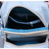 A light gray linen backpack is shown open, revealing a teal lining and decorative silver embroidery.